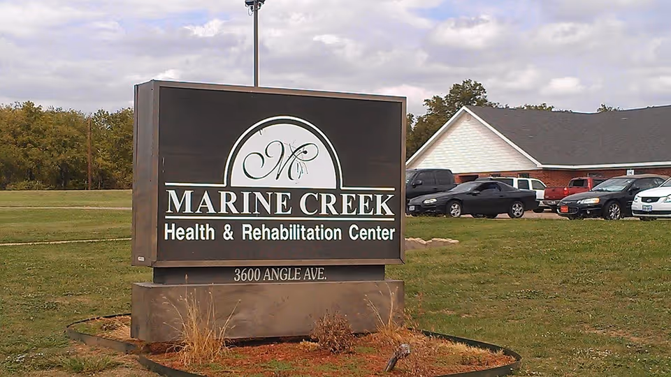 Outdoor view of a large sign for Marine Creek Health & Rehabilitation Center located at 3600 Angle Ave, with a parking lot and building in the background under a partly cloudy sky.