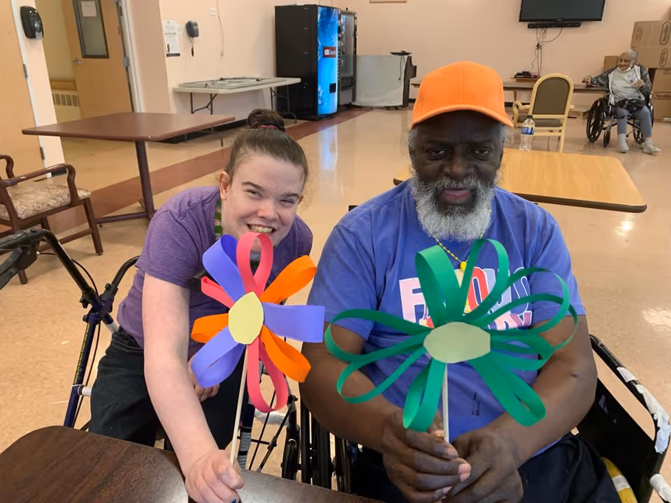 Two people sitting in wheelchairs inside a common room, smiling and holding colorful paper flowers. One person is wearing a purple shirt and the other is wearing a blue shirt and an orange cap. In the background, there is another person in a wheelchair and some tables and chairs.