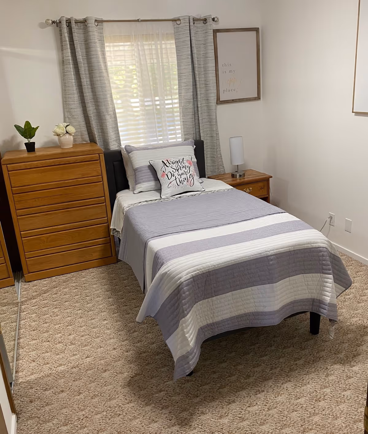 Neatly made single bed with gray-and-white striped bedding, a pillow, nightstand and dresser in a small, carpeted bedroom with a window and curtains.