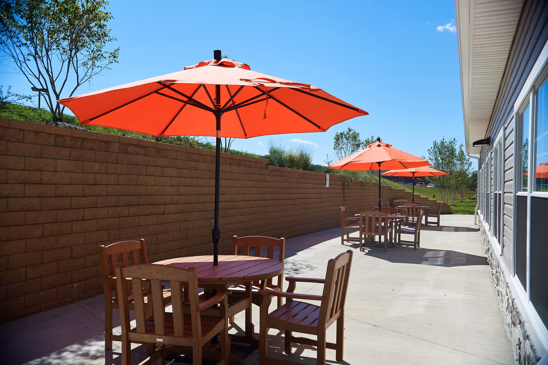 Outdoor patio with wooden tables and orange umbrellas along a building and retaining wall under a clear blue sky.