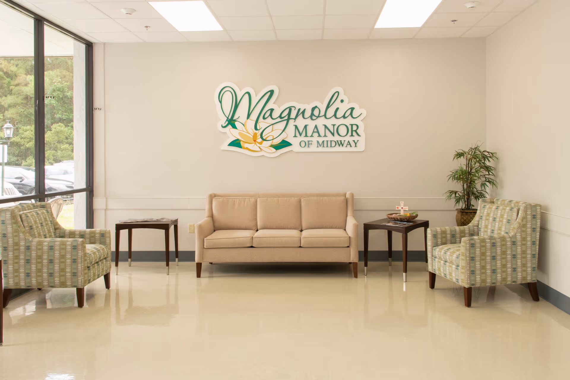 A waiting area with a beige sofa centered against a light-colored wall, flanked by two patterned armchairs on either side. There are two small dark wooden side tables next to the sofa, one with a decorative bowl and a small cross. A potted plant is placed in the corner to the right. Above the sofa is a sign that reads 'Magnolia Manor of Midway'. Large windows on the left side allow natural light to enter the room.