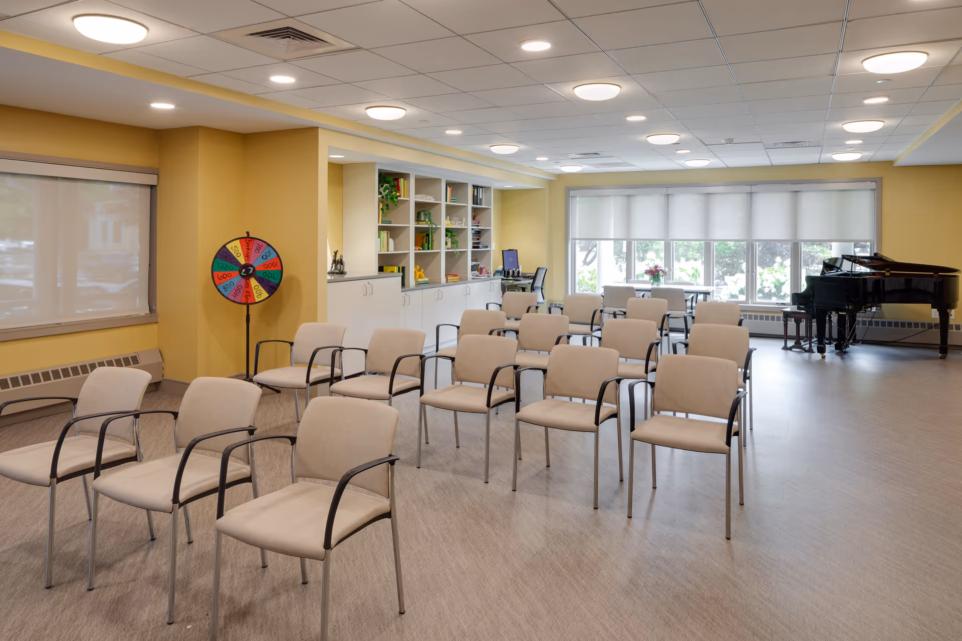 A spacious room with rows of beige chairs arranged facing forward, a colorful prize wheel on a stand near a yellow wall, built-in white shelves with books and decorative items, a computer workstation, large windows with white blinds, and a black grand piano in the corner.