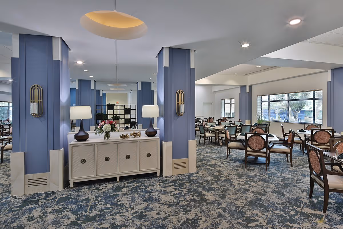 Spacious dining room with multiple tables and chairs arranged neatly. The room features blue accent pillars, a patterned carpet, large windows allowing natural light, and a white sideboard with a lamp and flower arrangement.