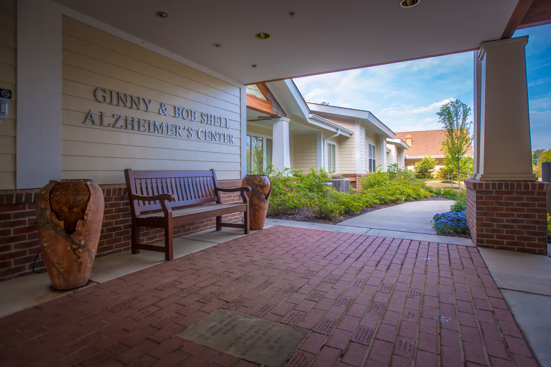 Covered entrance area of the Ginny & Bob Shell Alzheimer's Center at Parkway Village, featuring a wooden bench, two large decorative vases, brick flooring with engraved bricks, and a pathway leading to the building surrounded by greenery and blue sky.