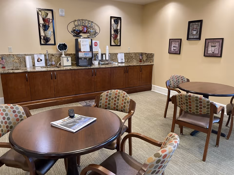 A cozy common area with round wooden tables and chairs featuring colorful polka dot upholstery. Against the wall is a countertop with cabinets below, a coffee machine, paper cups, and decorative coffee-themed wall art above.