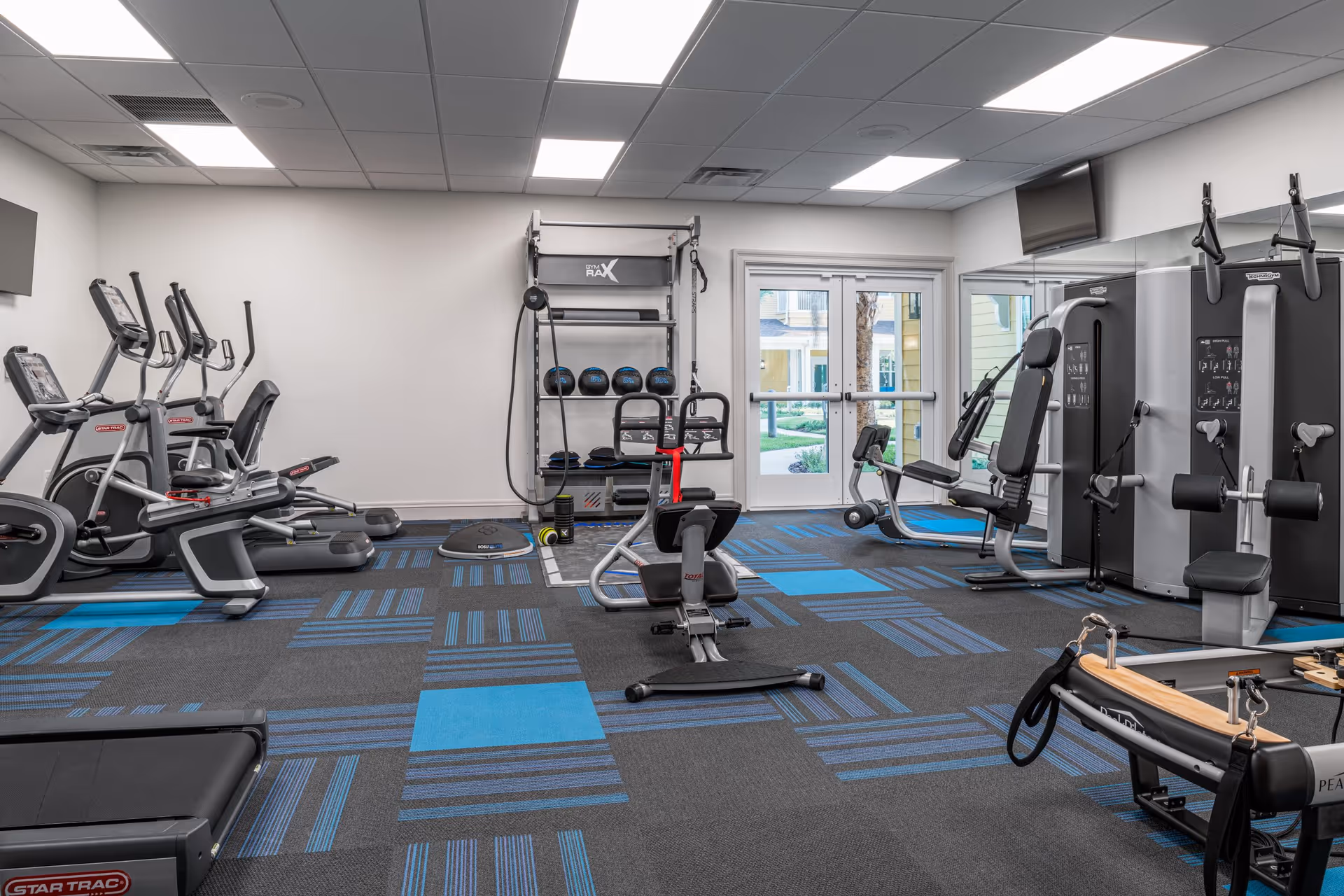 A well-lit fitness room with various exercise equipment including treadmills, elliptical machines, weight machines, a rowing machine, and a rack with medicine balls and other fitness accessories. The room has a patterned carpet with blue and gray squares, white walls, and a ceiling with recessed lighting. There are double glass doors leading outside, and a large mirror on one wall.