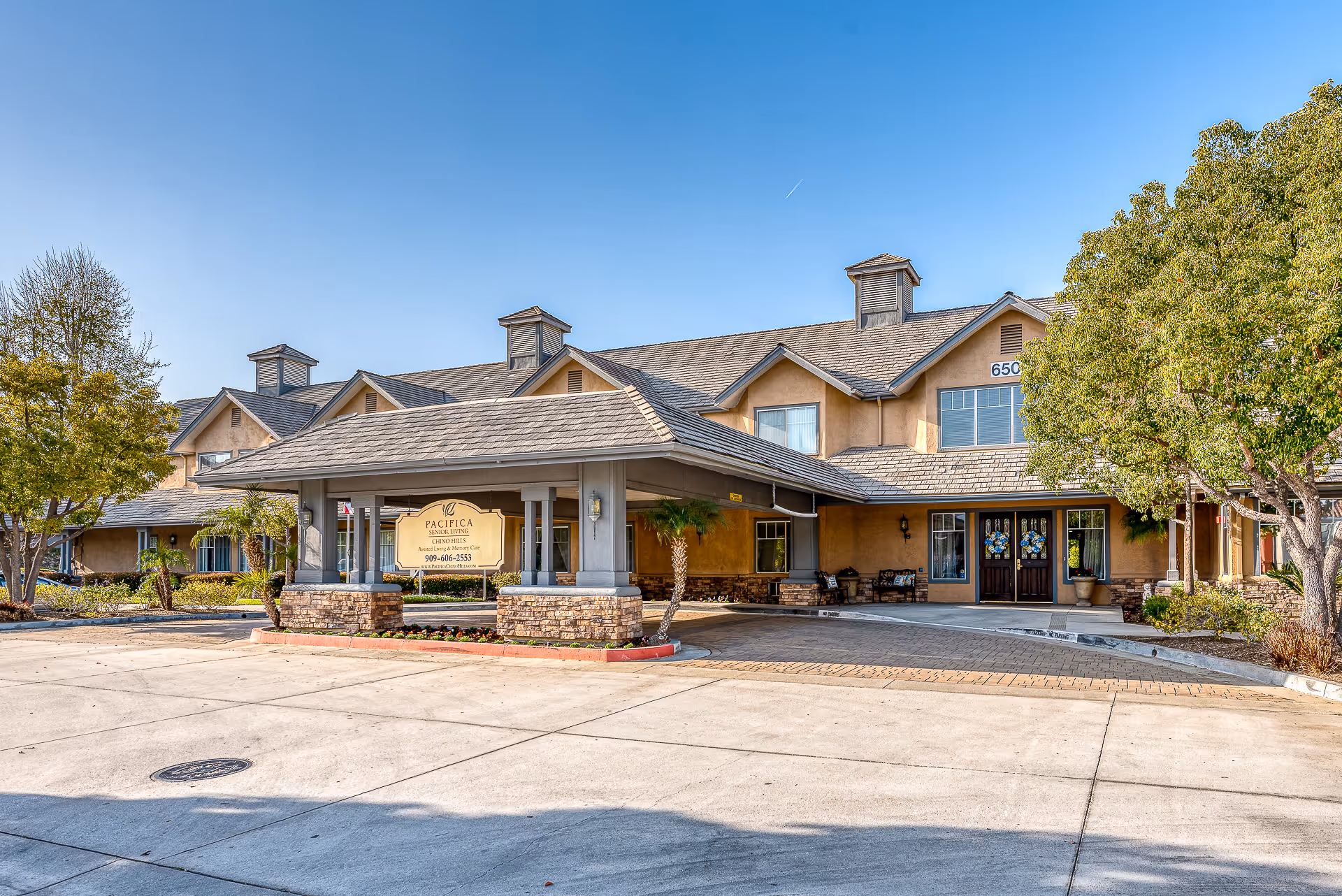 Exterior view of Pacifica Senior Living Chino Hills building with a covered entrance, stone pillars, trees, and clear blue sky.