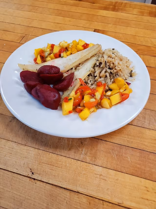 A white plate on a wooden table with a meal consisting of cooked white fish fillets, wild rice, cooked beets, and a colorful mango and red pepper salsa.