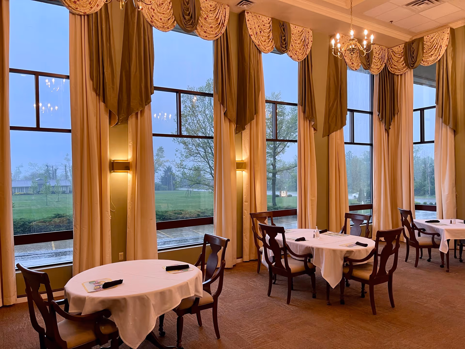 A formal dining room with round tables and chairs set near large draped windows overlooking a lawn.