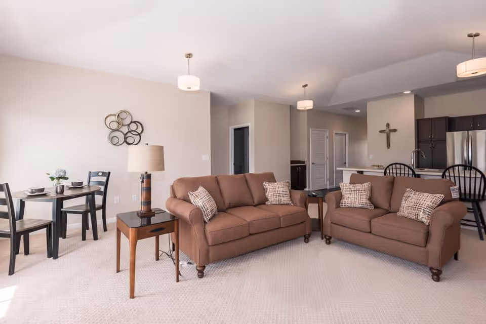 A bright and spacious living area featuring two brown sofas with patterned cushions, a wooden side table with a lamp, a small dining table with four chairs set with dishes, and a kitchen area with a breakfast bar and black chairs. The walls are light-colored, and there is a decorative wall piece and a cross hanging on the walls.