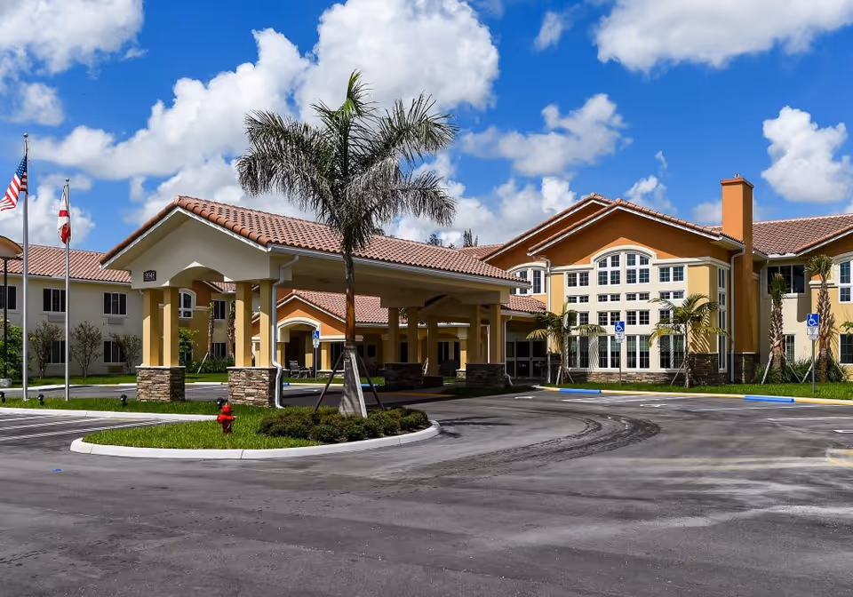 Front exterior of a Mediterranean-style senior living building with a covered entrance, palm tree, flags, and a parking lot under a blue sky.