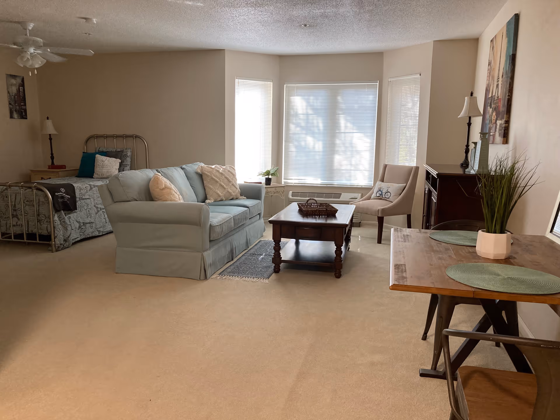 A bright and cozy studio apartment with a light blue sofa, wooden coffee table, beige armchair with a bicycle pillow, and a small dining table with two chairs and green placemats. In the background, there is a metal-framed bed with pillows and a nightstand with a lamp. Large windows with blinds allow natural light to fill the room.