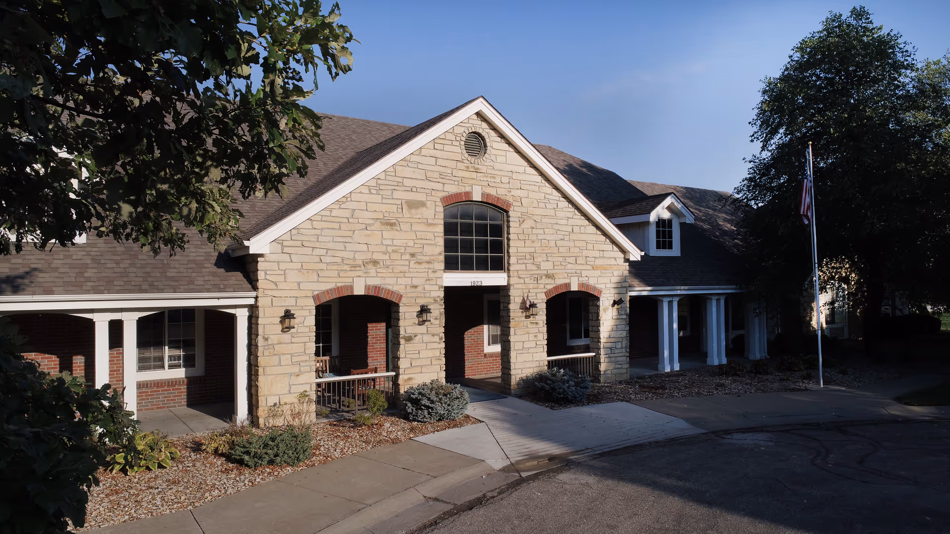 Front entrance of a stone-faced assisted living building with arched entryways, columns, landscaping, and an American flag.