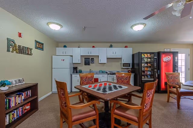 A common area in a senior living facility featuring a small kitchen with white cabinets, a refrigerator, microwave, and coffee maker. In the foreground, there is a square table with a checkerboard game set up and four wooden chairs with patterned cushions around it. To the right, there are vending machines including a Dr Pepper machine. On the left wall, there is a bookshelf filled with books and a decorative sign that says 'FAMILY'. The room has beige walls and carpeted floor, with ceiling lights and a ceiling fan visible.