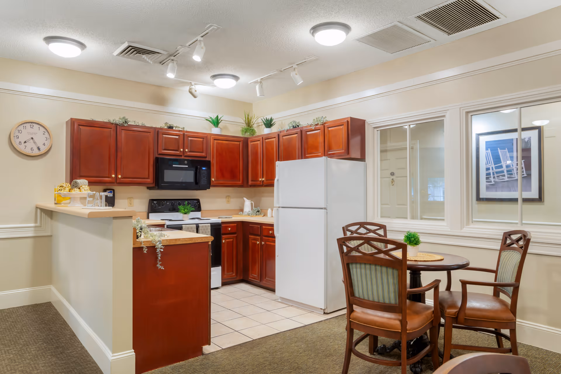 A cozy kitchen area with wooden cabinets, a white refrigerator, a black microwave, and a stove. There is a small round dining table with three wooden chairs with green cushions. The walls are light-colored with a clock and framed picture visible. The floor is a combination of tile in the kitchen area and carpet in the dining area. Ceiling lights and track lighting illuminate the space.