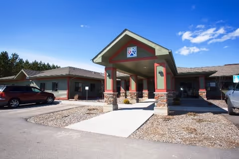 Exterior view of Babbitt Carefree Living facility showing the main entrance with a covered driveway supported by stone and wood pillars. The building has a green and red color scheme with a clear blue sky above. Cars are parked on either side of the entrance.