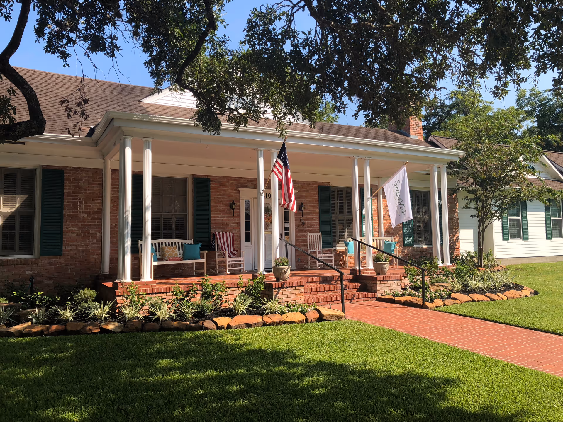 Front exterior view of a brick and white senior living facility building with a covered porch featuring white columns, rocking chairs, and two flags, one American and one with the Seasons logo. The building is surrounded by a well-maintained lawn, plants, and trees under a clear blue sky.