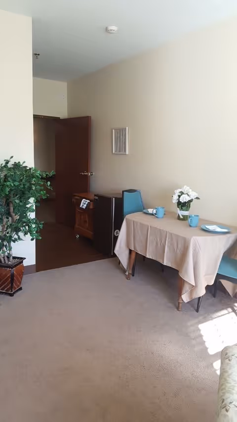 A small dining area in an assisted living facility with a table covered by a beige tablecloth, two blue chairs, a vase with white flowers, two blue mugs, and a plate. There is a potted plant on the left side near an open wooden door, and a small cabinet with a mini fridge next to the table. The room has beige walls and carpeted floor with sunlight coming through a window.