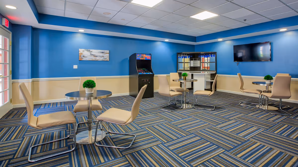 Bright communal lounge with blue walls, small round tables and beige chairs, a wall-mounted TV and an arcade machine.