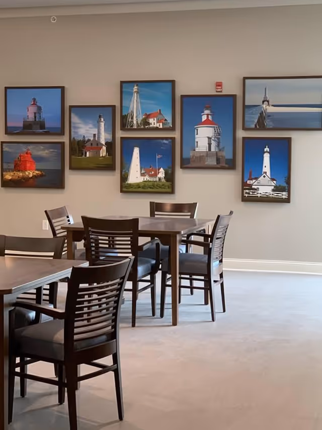A dining area with wooden tables and chairs arranged on a light-colored floor. The wall behind the tables features a collection of framed photographs of various lighthouses against blue skies.