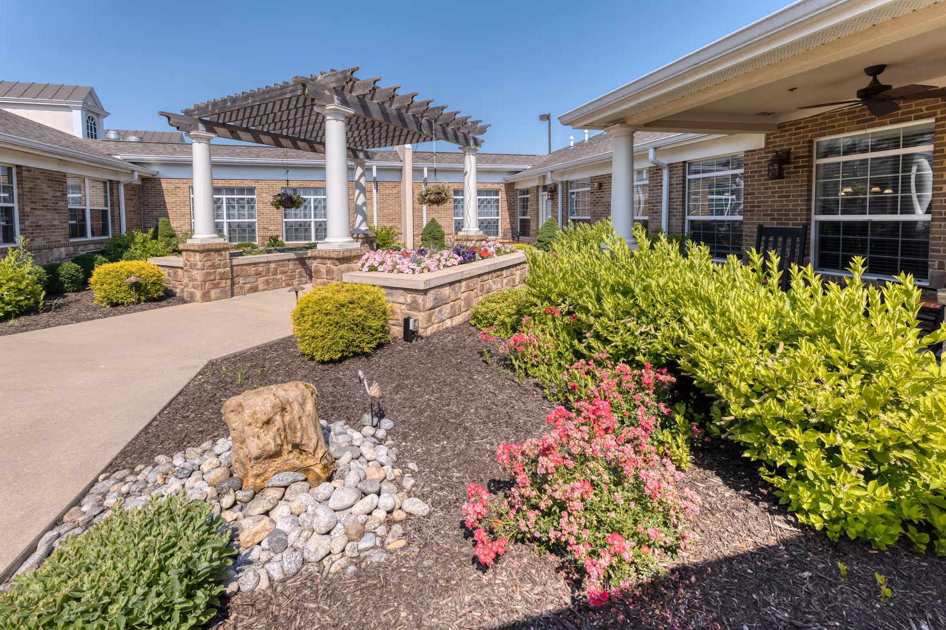 Outdoor courtyard area at Addington Place of Shoal Creek featuring a pergola with white columns, flower beds with pink and green plants, a rock garden, and brick building walls with large windows in the background under a clear blue sky.