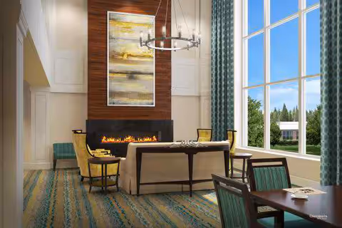 Bright communal living room with high ceilings, a modern fireplace, seating and large windows overlooking greenery.