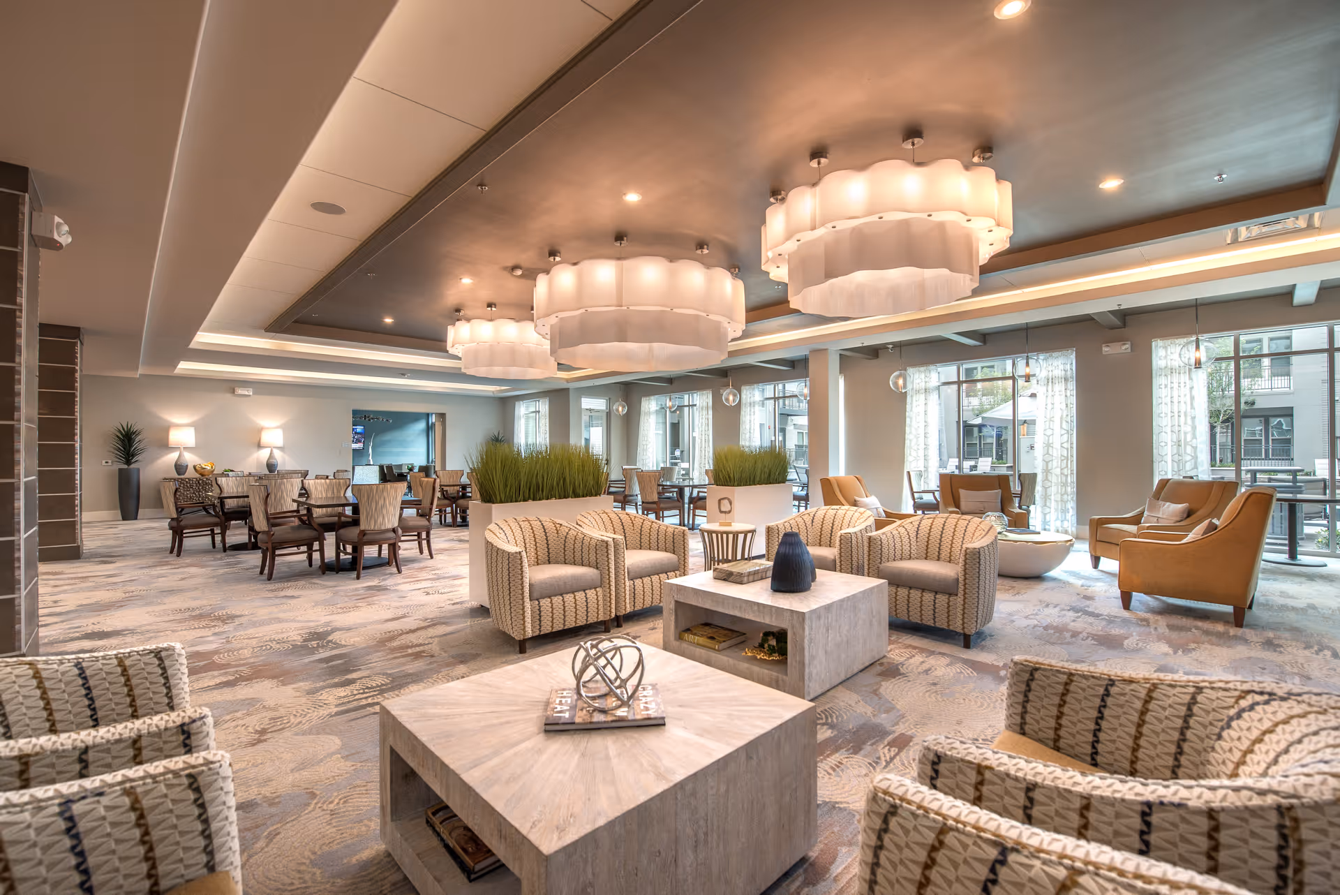 Spacious and well-lit senior living facility common area with modern decor, featuring multiple seating arrangements including patterned armchairs, wooden dining tables with chairs, large square coffee tables with decorative items, and large windows with sheer curtains allowing natural light to fill the room. The ceiling has recessed lighting and large, elegant pendant lights.