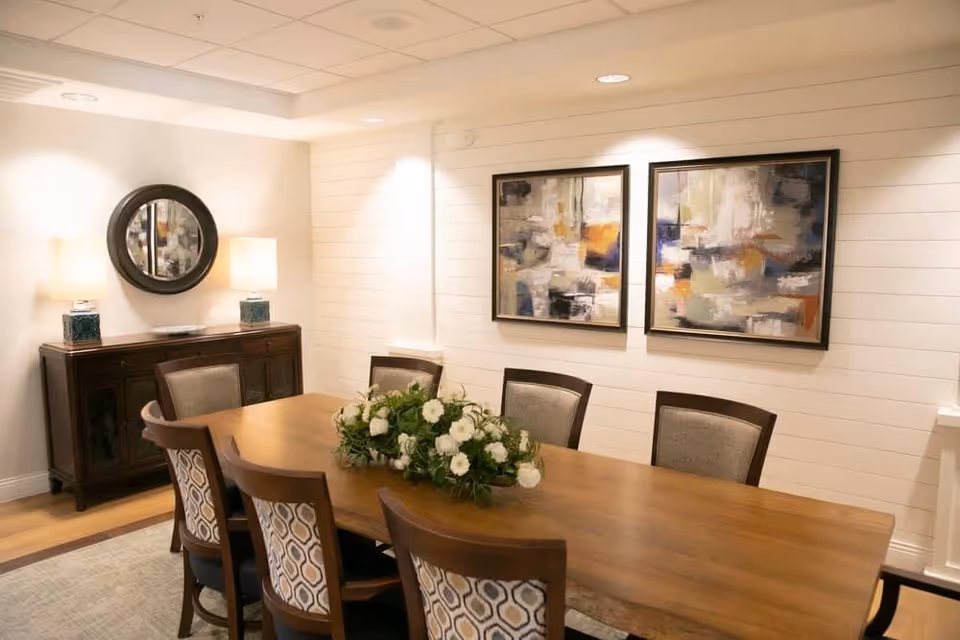 A dining room with a long wooden table surrounded by eight chairs, each with patterned upholstery on the backrests. A floral centerpiece with white flowers and greenery is placed on the table. On the wall behind the table, there are two abstract paintings with a mix of colors including white, gray, orange, and blue. To the left, there is a dark wooden sideboard with two table lamps and a round mirror hanging above it. The room has light-colored walls with horizontal paneling and a light-colored carpet on the floor.