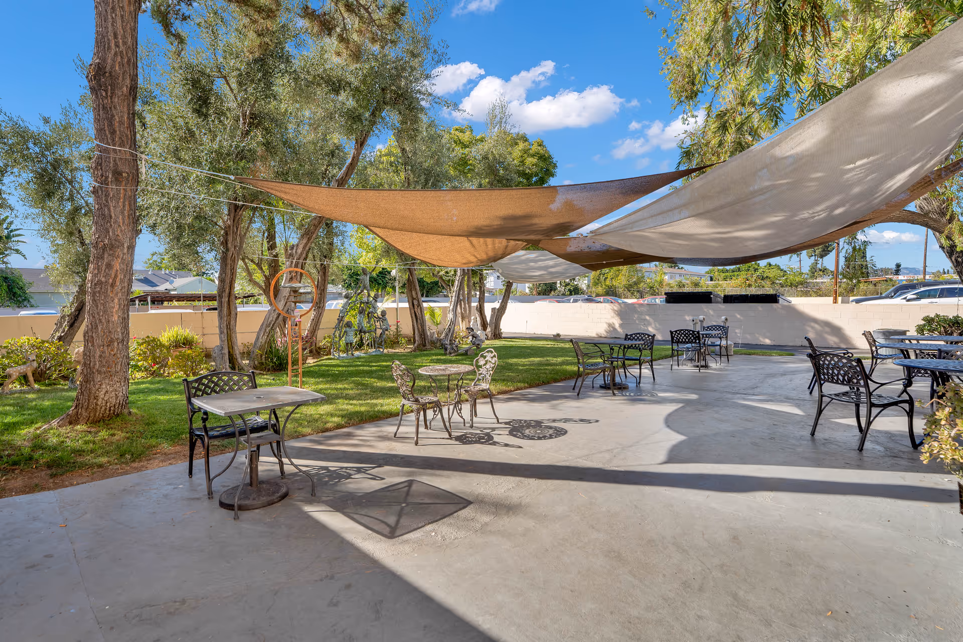 Shaded outdoor patio with metal tables and chairs under fabric sails next to a grassy area and trees.