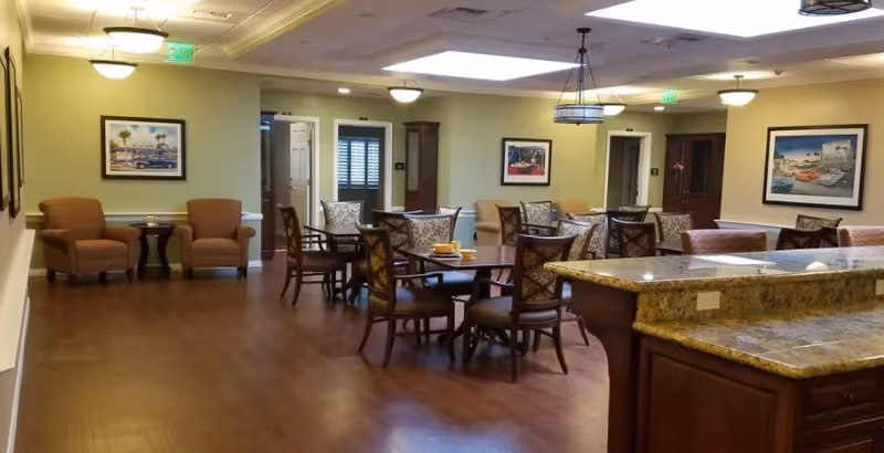 A spacious dining area in a senior living facility with multiple round tables and chairs arranged for seating. The room features wooden flooring, beige walls adorned with framed artwork, and comfortable armchairs along the walls. A granite countertop is visible in the foreground, and ceiling lights provide warm illumination.