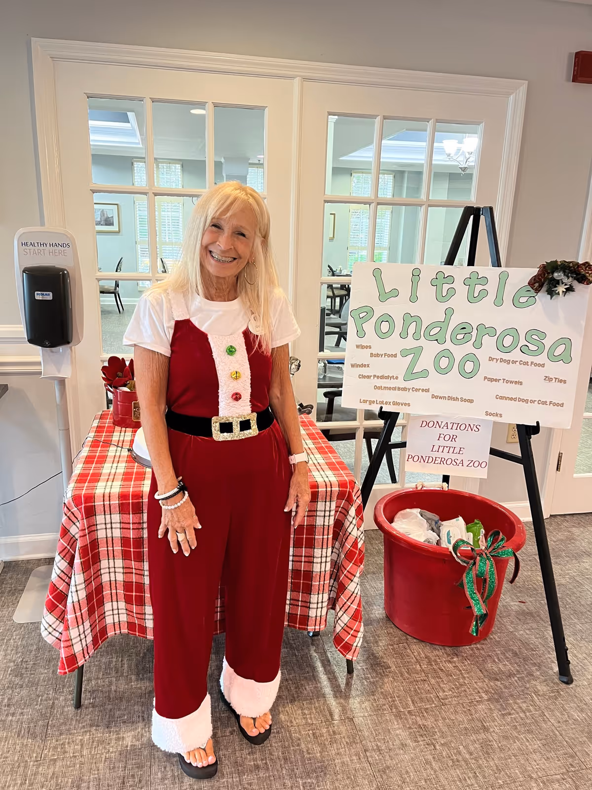 A smiling elderly woman dressed in a festive red and white outfit resembling a Santa suit stands indoors next to a table covered with a red and white plaid tablecloth. Behind her is a sign on an easel that reads 'Little Ponderosa Zoo' and lists various donation items. There is also a large red bucket decorated with a green and red ribbon for collecting donations. The setting appears to be a community or senior living facility with glass-paned doors and a hand sanitizer dispenser on the wall.