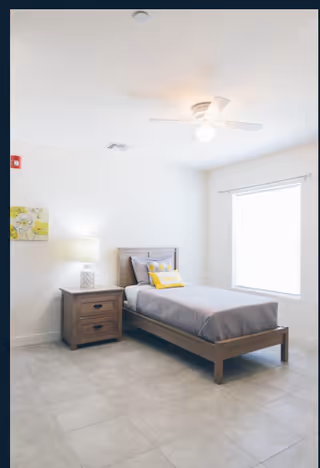 A simple, bright bedroom with a single wooden bed made with gray bedding and a yellow accent pillow. Next to the bed is a wooden nightstand with a lamp. The room has a large window with white curtains and a ceiling fan with a light.
