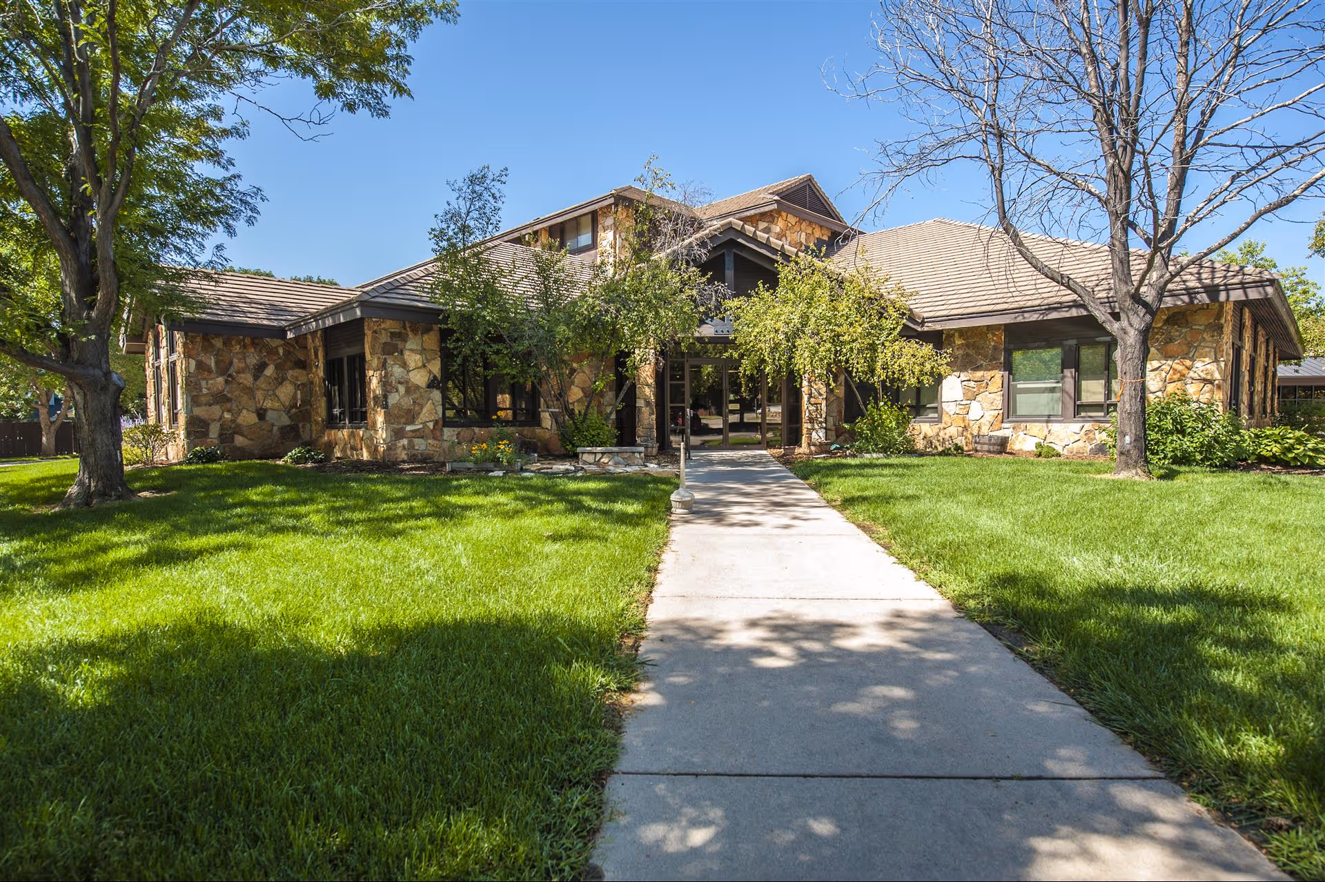 Front exterior view of a single-story assisted living facility building with stone facade, surrounded by green grass, trees, and a concrete walkway leading to the entrance under a clear blue sky.