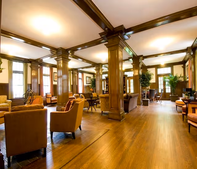 Spacious senior living facility common area with wooden floors, large wooden columns, multiple seating arrangements including armchairs and sofas, large windows letting in natural light, and several potted plants.