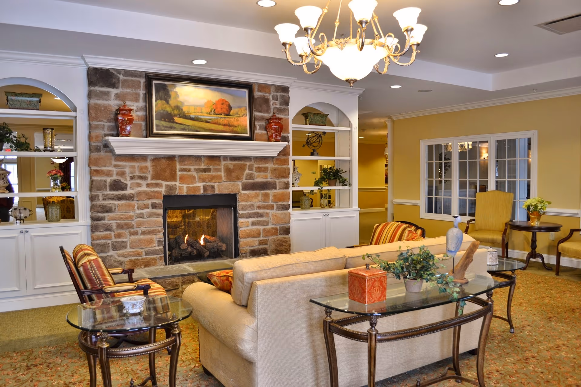 A cozy living room with a stone fireplace in the center, a beige sofa facing the fireplace, two striped armchairs, a glass coffee table with decorative items, built-in white shelves on either side of the fireplace, yellow walls, and a chandelier hanging from the ceiling.