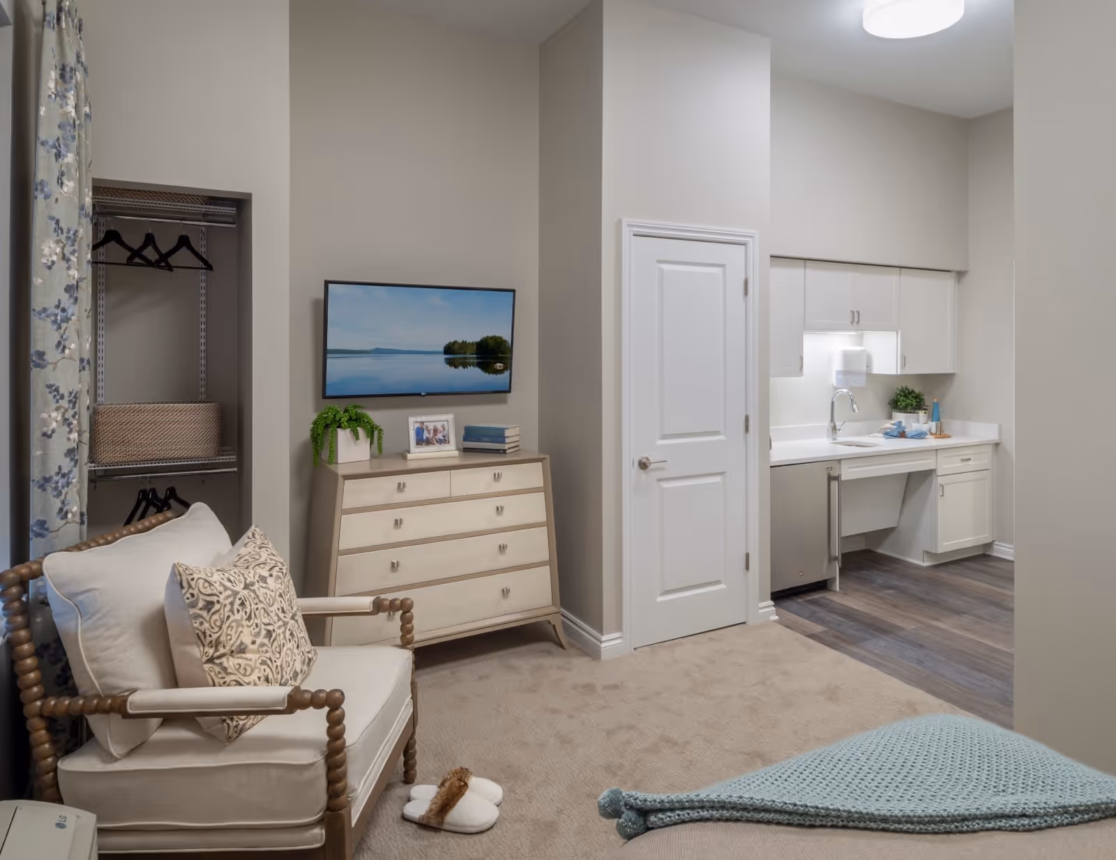 A cozy senior living room area with a cushioned armchair and decorative pillow, a dresser with a plant, photo frame, and books, a wall-mounted TV displaying a serene lake scene, an open closet with hangers and a basket, and a small kitchenette with white cabinets, a sink, and countertop. The room has beige walls and carpet, with wood flooring in the kitchenette area.
