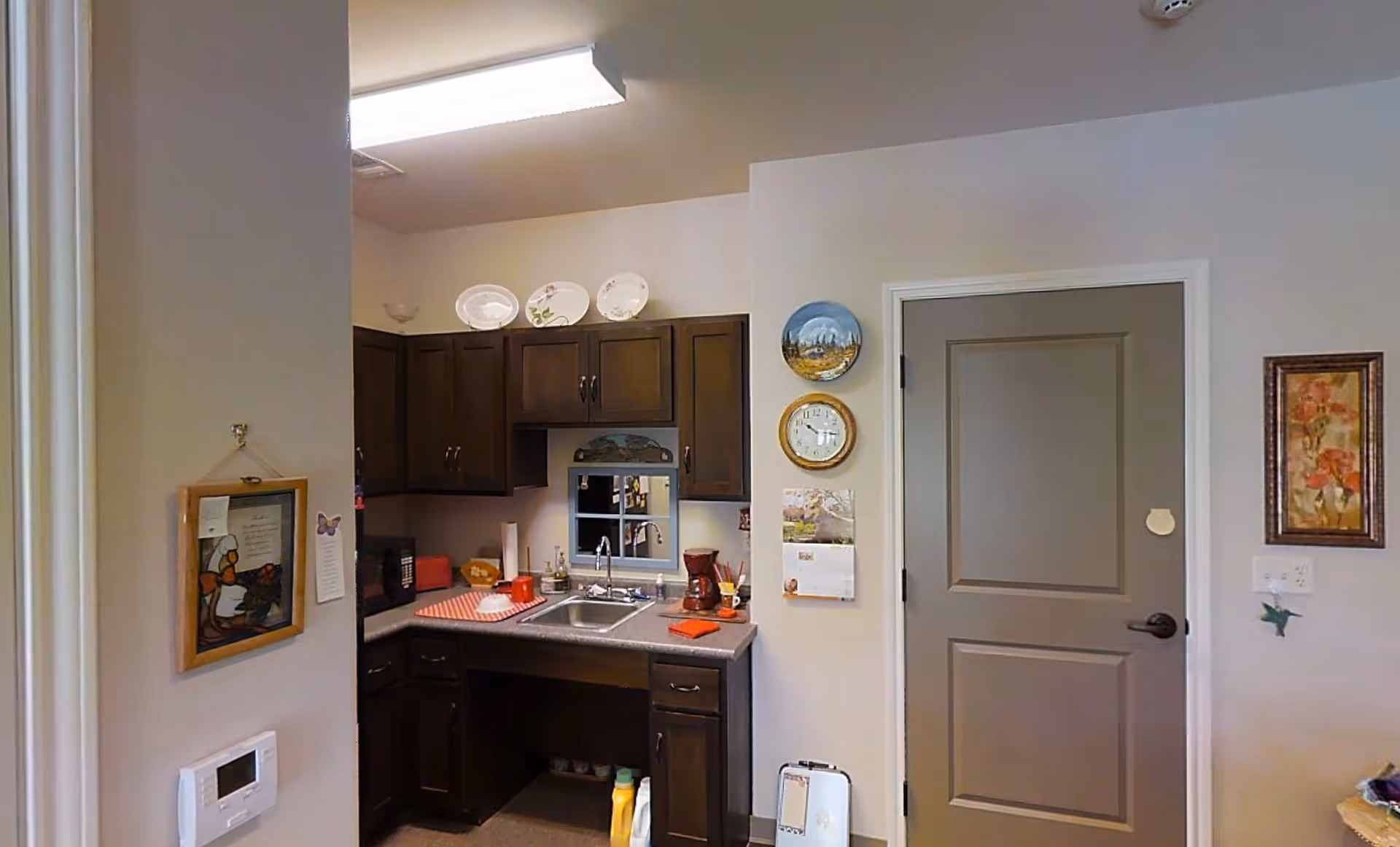 A small kitchen area with dark wooden cabinets, a countertop with a sink, a coffee maker, and various kitchen items. Decorative plates are displayed above the cabinets, and a clock and calendar hang on the adjacent wall. A closed gray door is visible to the right, and framed artwork is hung on the walls.