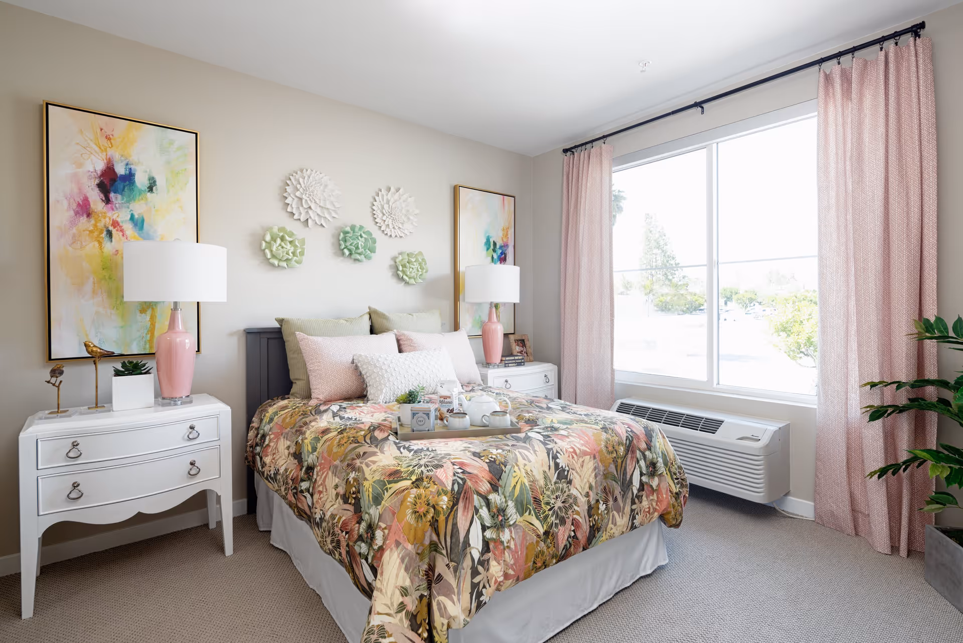 A bright and cozy bedroom with a floral patterned bedspread on a bed, multiple pillows, and a tray with tea set on top. The room features two white nightstands with pink lamps, colorful abstract paintings on the wall, decorative wall art above the bed, a large window with pink curtains, a wall-mounted air conditioning unit below the window, and a green potted plant in the corner.