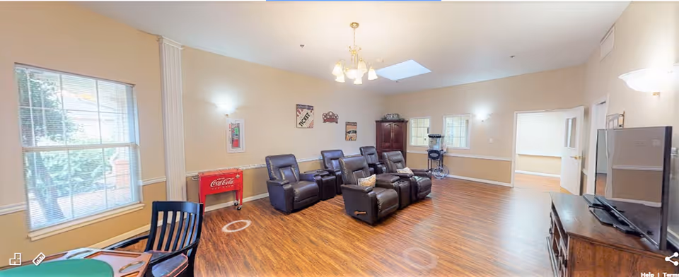 A spacious living room in an assisted living community featuring six black leather recliners arranged in two rows facing a large flat-screen TV on a wooden stand. The room has wood flooring, beige walls with white trim, two windows letting in natural light, a skylight, and wall-mounted lights. There is a red Coca-Cola cooler against one wall and a small table with chairs in the foreground.