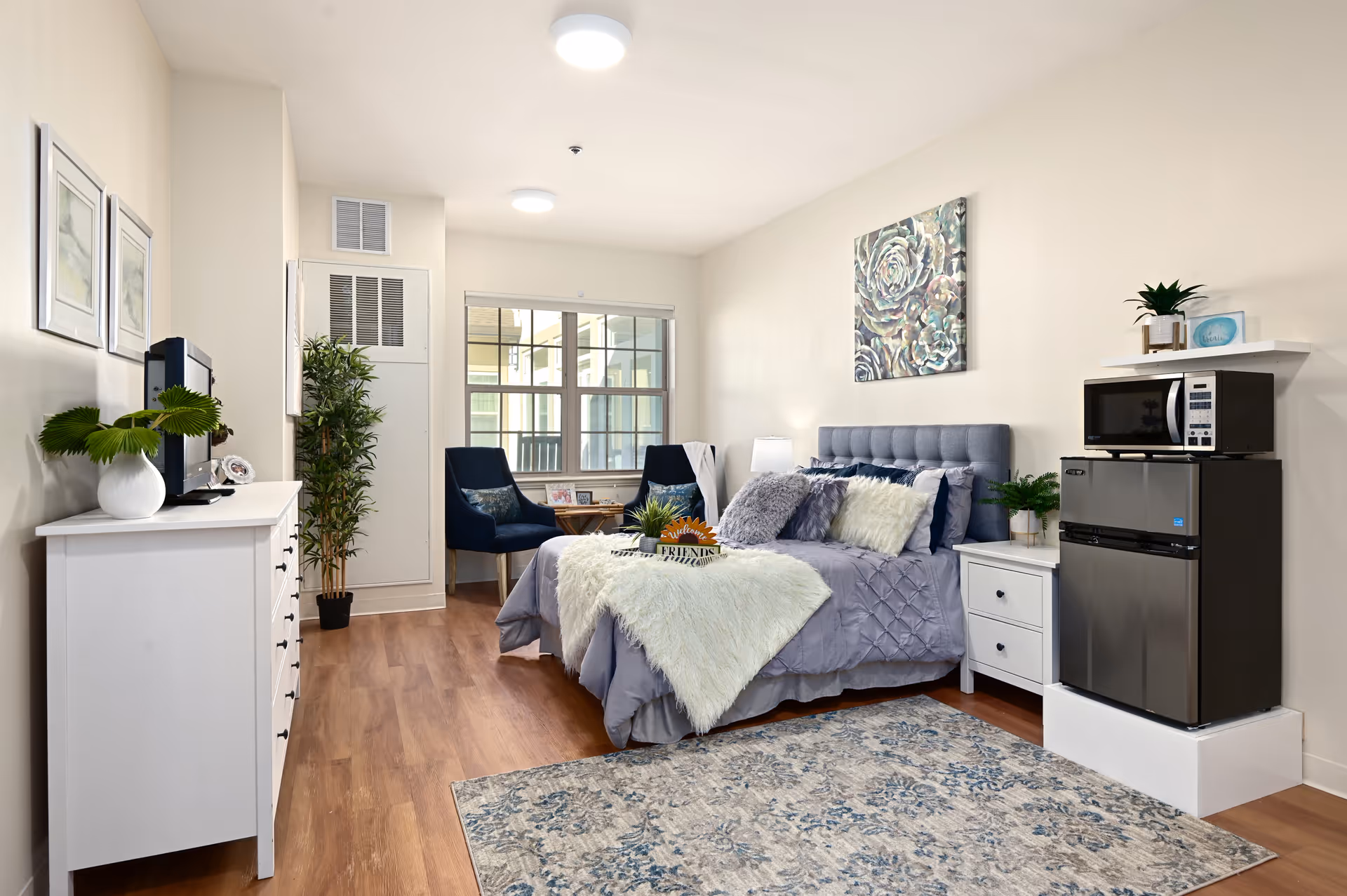 A cozy bedroom in a senior living facility featuring a neatly made bed with gray and white bedding and multiple pillows. There is a white nightstand with a small plant and a lamp beside the bed. A small refrigerator and microwave are placed on a white stand to the right. Two blue chairs and a small round table are positioned near a window at the back. The room has light-colored walls, wooden flooring, a patterned area rug, and decorative artwork on the walls.
