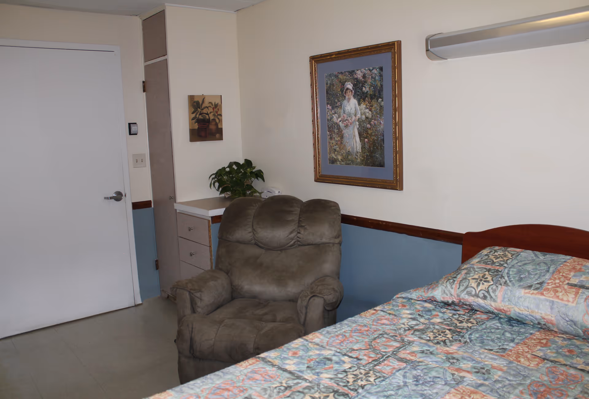 A room with a bed covered in a colorful patterned quilt, a brown recliner chair, a small counter with drawers, a potted plant, and two framed paintings on the wall. The walls are painted beige with a blue lower half and there is a white door on the left side.