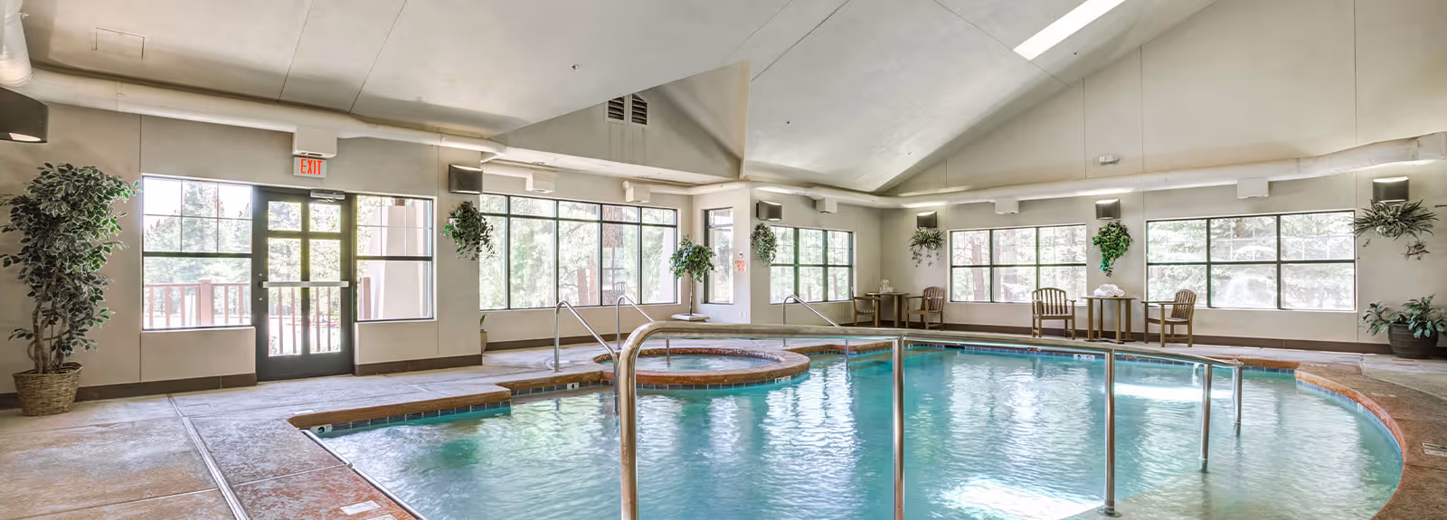Indoor swimming pool area with a hot tub, surrounded by large windows letting in natural light. There are several potted plants and small seating areas with wooden chairs and tables around the pool. The ceiling is vaulted with exposed ductwork and there is an exit door on the left side.