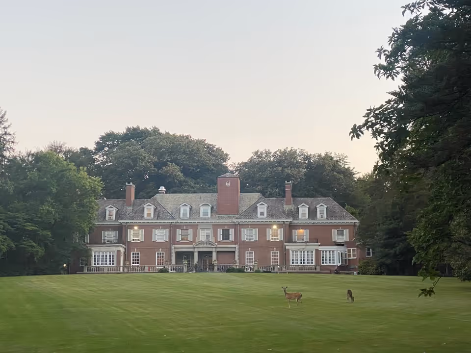 Large brick manor house with an expansive lawn and two deer grazing in front, framed by trees.