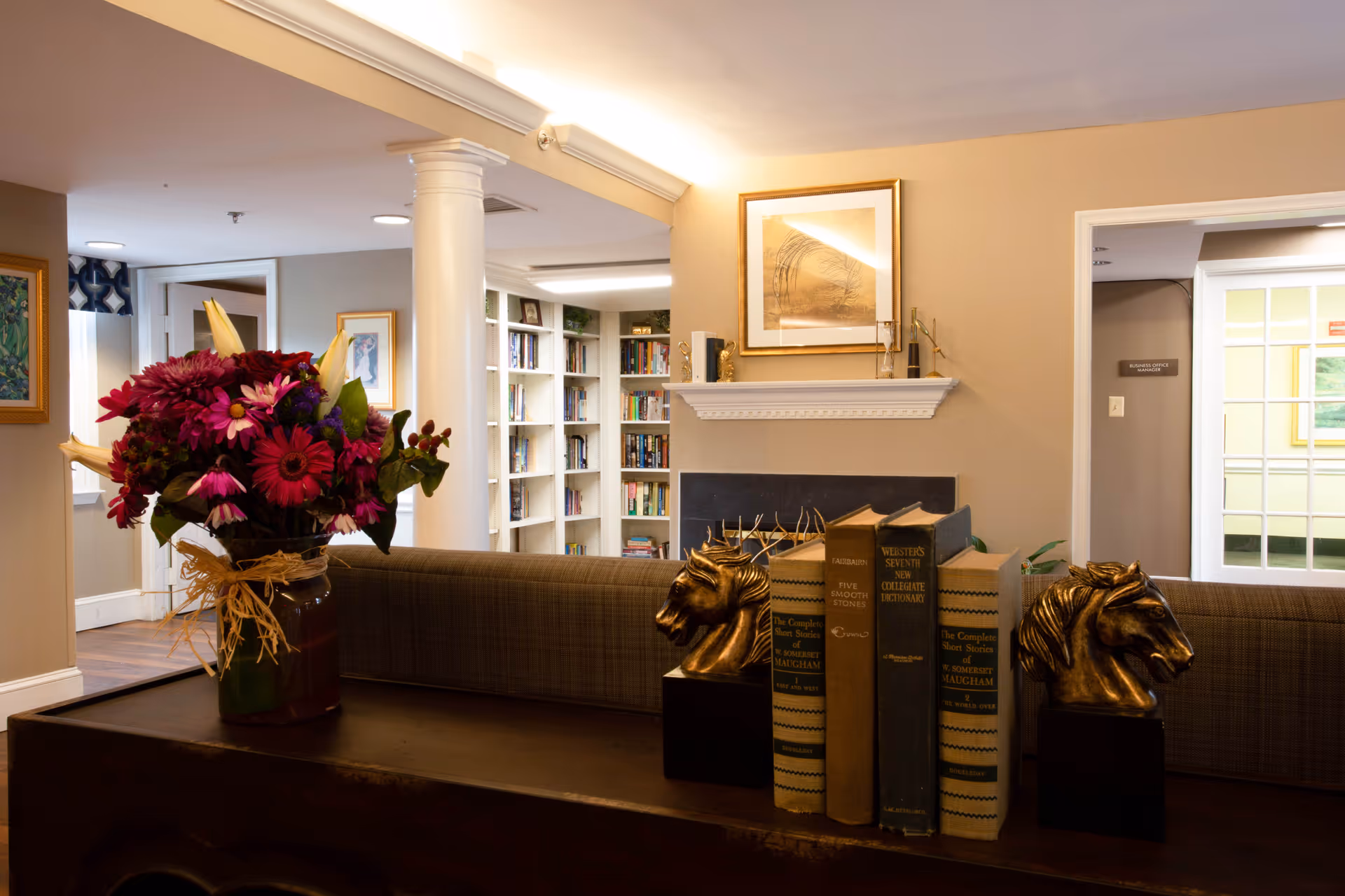 Interior view of a cozy living room area with a dark wooden table in the foreground holding a vase of vibrant flowers and several books supported by horse head bookends. In the background, there is a fireplace with a framed picture above it, built-in bookshelves filled with books, and doorways leading to other rooms.