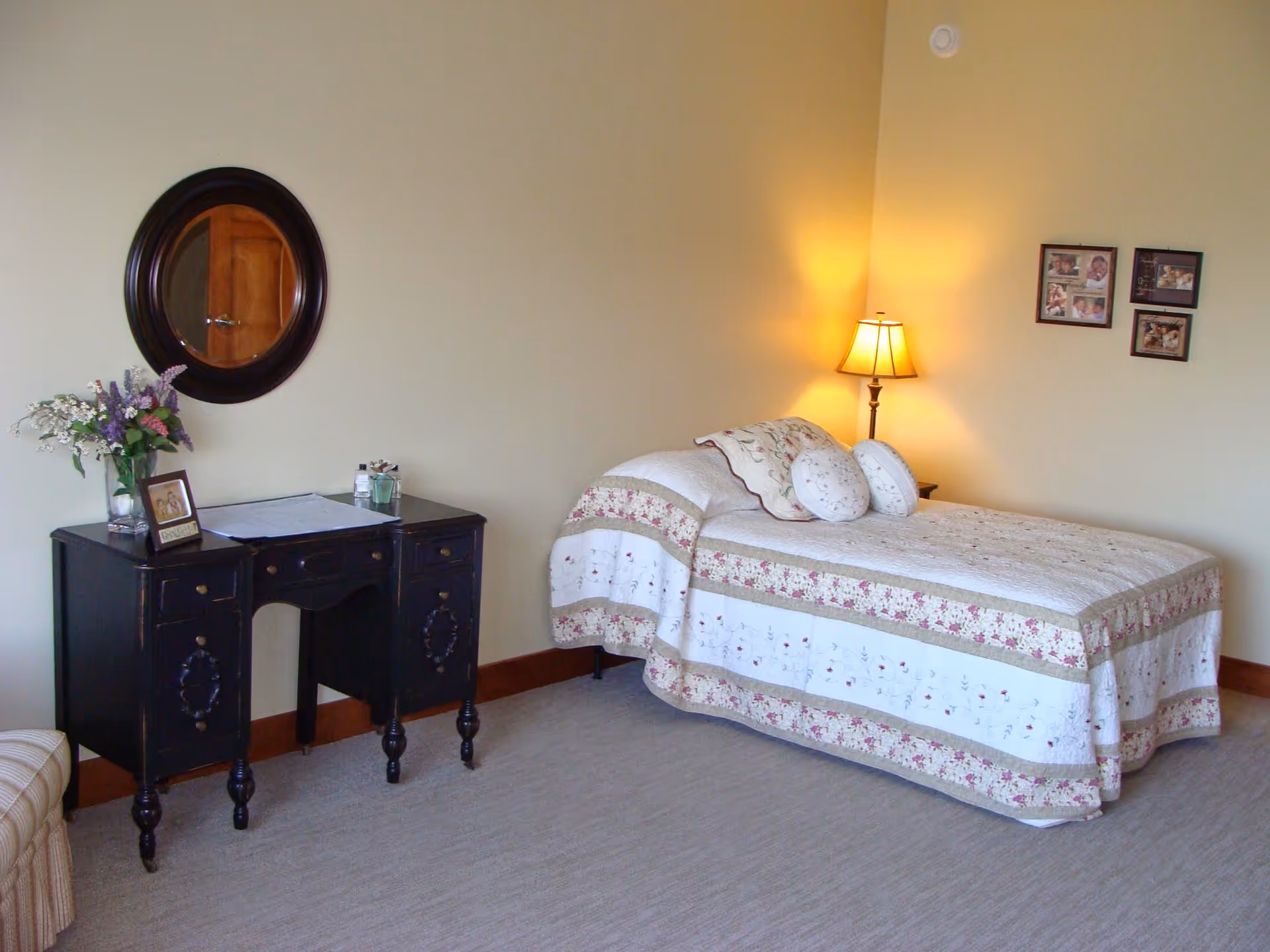 A cozy bedroom with a single bed covered in a floral quilt and matching pillows. A floor lamp with a warm light stands next to the bed. On the wall above the bed are four framed pictures. To the left of the bed is a black vintage-style desk with a vase of flowers, a framed photo, and some small items on top. A round mirror hangs on the wall above the desk. The room has beige walls and carpeted flooring.