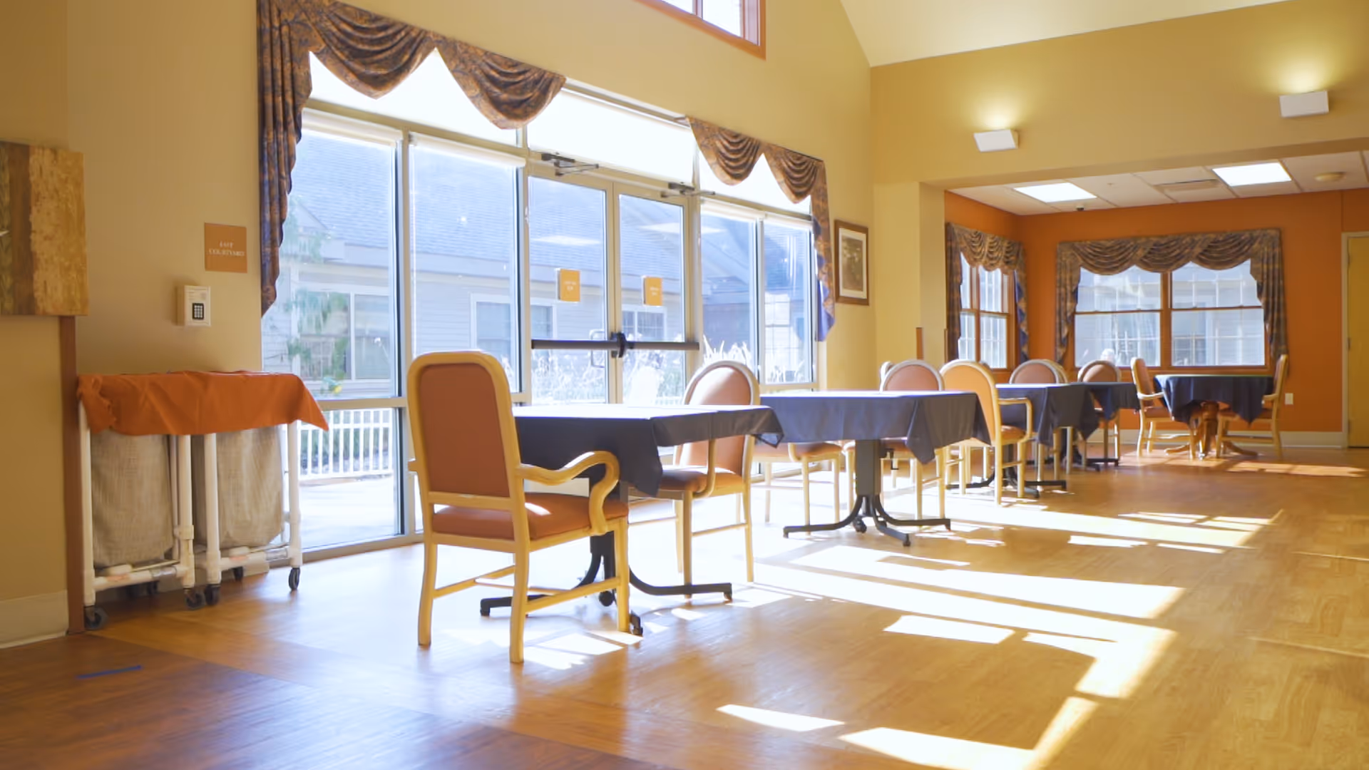 A bright dining area with several tables covered in dark tablecloths and surrounded by wooden chairs with orange cushions. Large windows with decorative valances allow natural light to fill the room, casting shadows on the wooden floor. The walls are painted in warm tones, and there is a laundry cart with an orange cover on the left side near the windows.