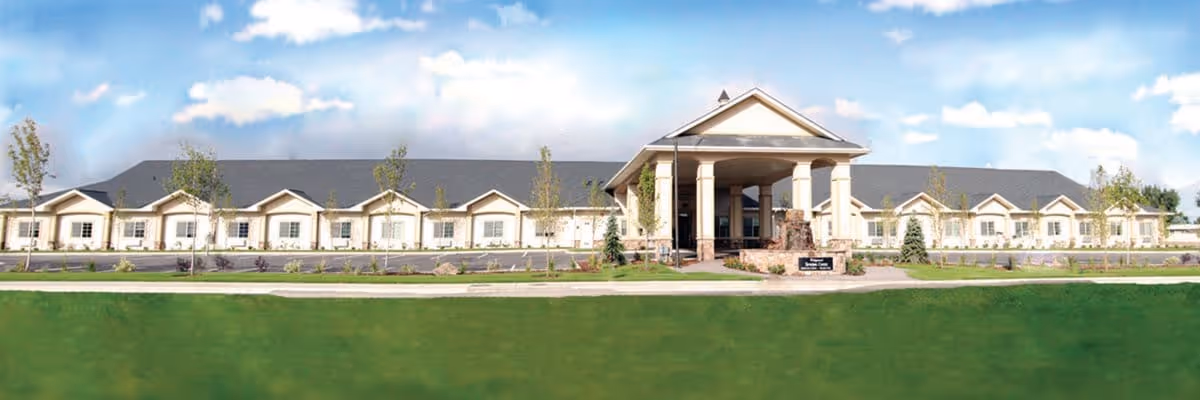 Wide exterior view of a single-story senior living facility building with a large covered entrance supported by columns, surrounded by a well-maintained lawn and young trees under a partly cloudy sky.