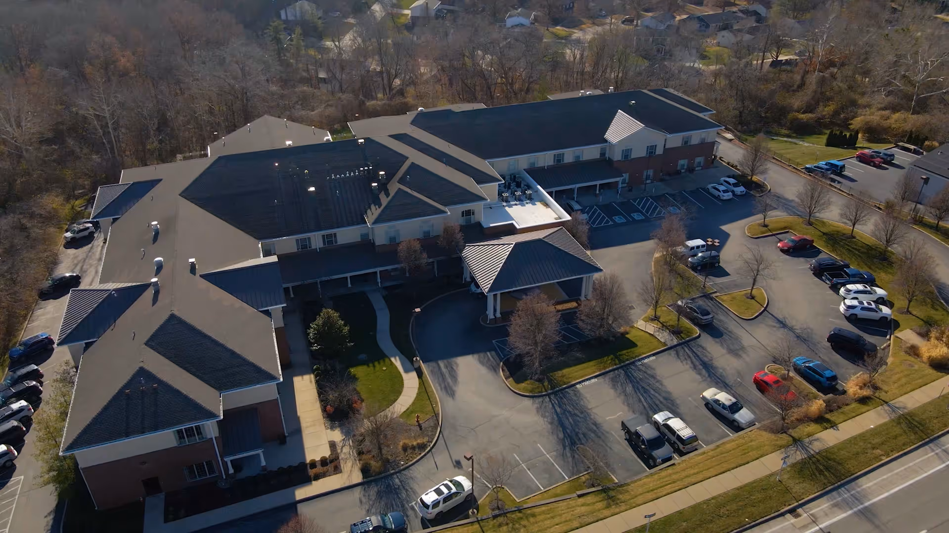 Aerial view of a senior living facility named Quarters At Des Peres showing a large building with multiple wings, a covered entrance, surrounding parking lots with several parked cars, and some trees and greenery around the property.