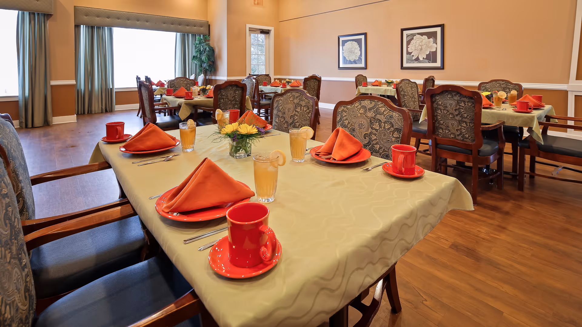 A dining room with several tables covered in light green tablecloths, each set with red plates, red cups, orange folded napkins, silverware, and glasses of iced tea garnished with lemon slices. The room has wooden floors, beige walls with white trim, framed floral artwork, and windows with green curtains.