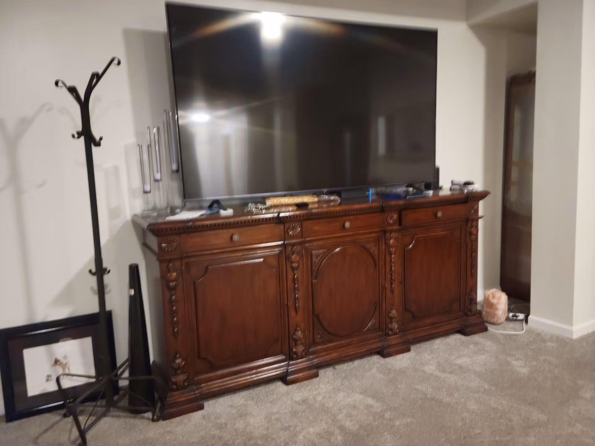 A large flat-screen TV sits on an ornate wooden sideboard in a carpeted living room with a coat rack and a framed picture leaning against the wall.